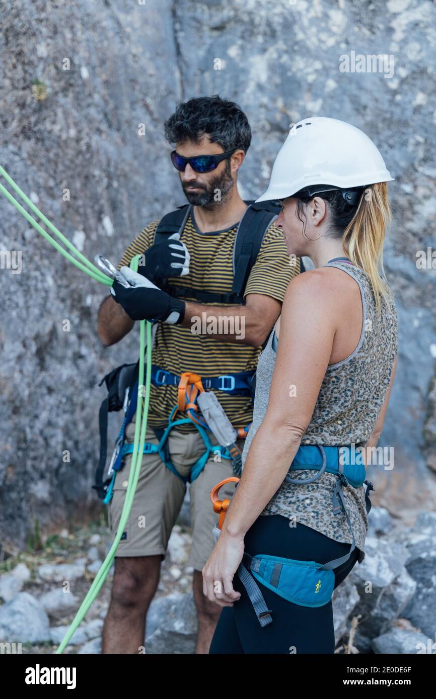 Fearless active couple in safety harnesses preparing gear for ascending rocky mountain slope while practicing climbing in nature Stock Photo