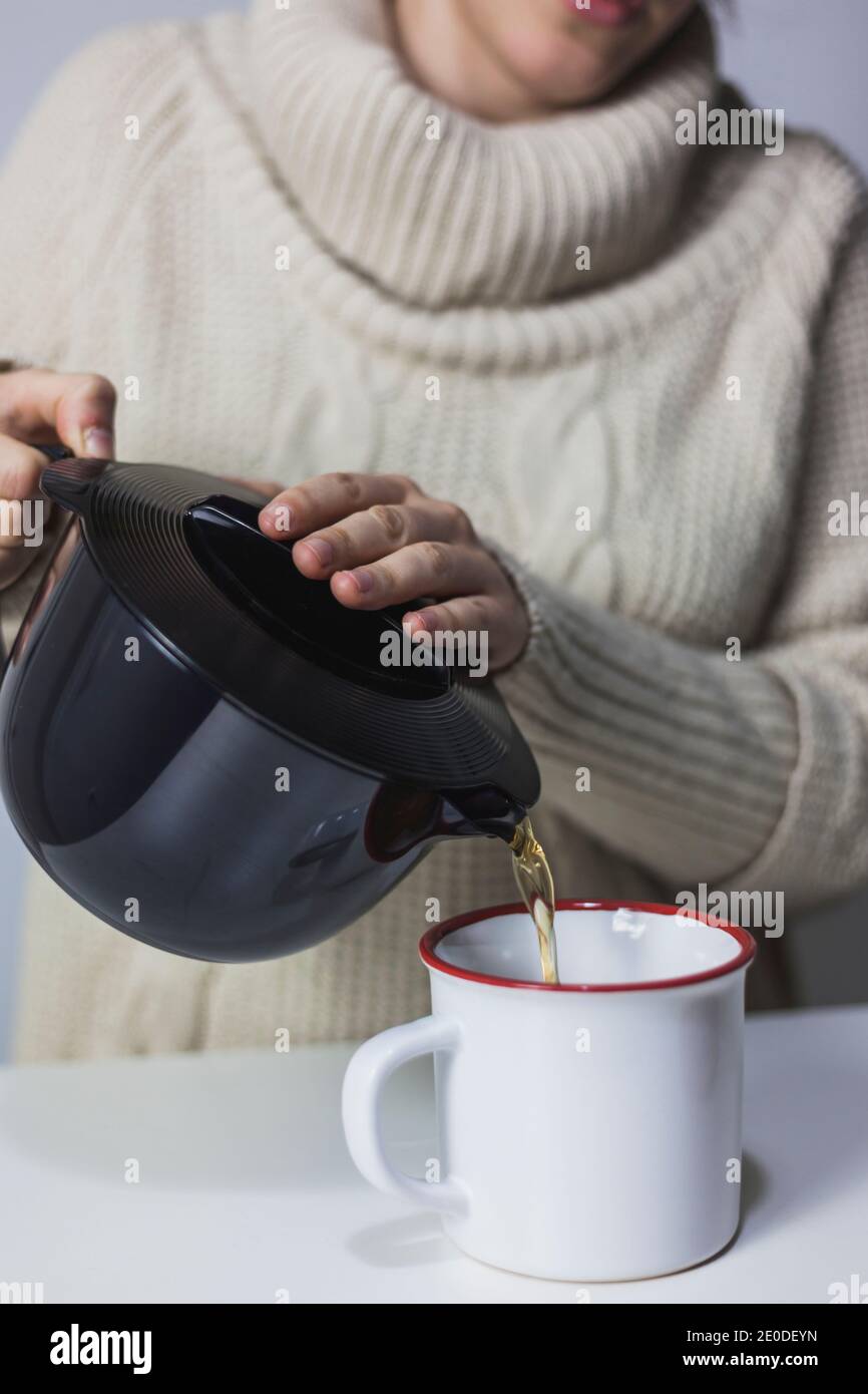 Unrecognizable crop female in knitted sweater pouring tasty aromatic tea from teapot in mug while enjoying weekend at home Stock Photo