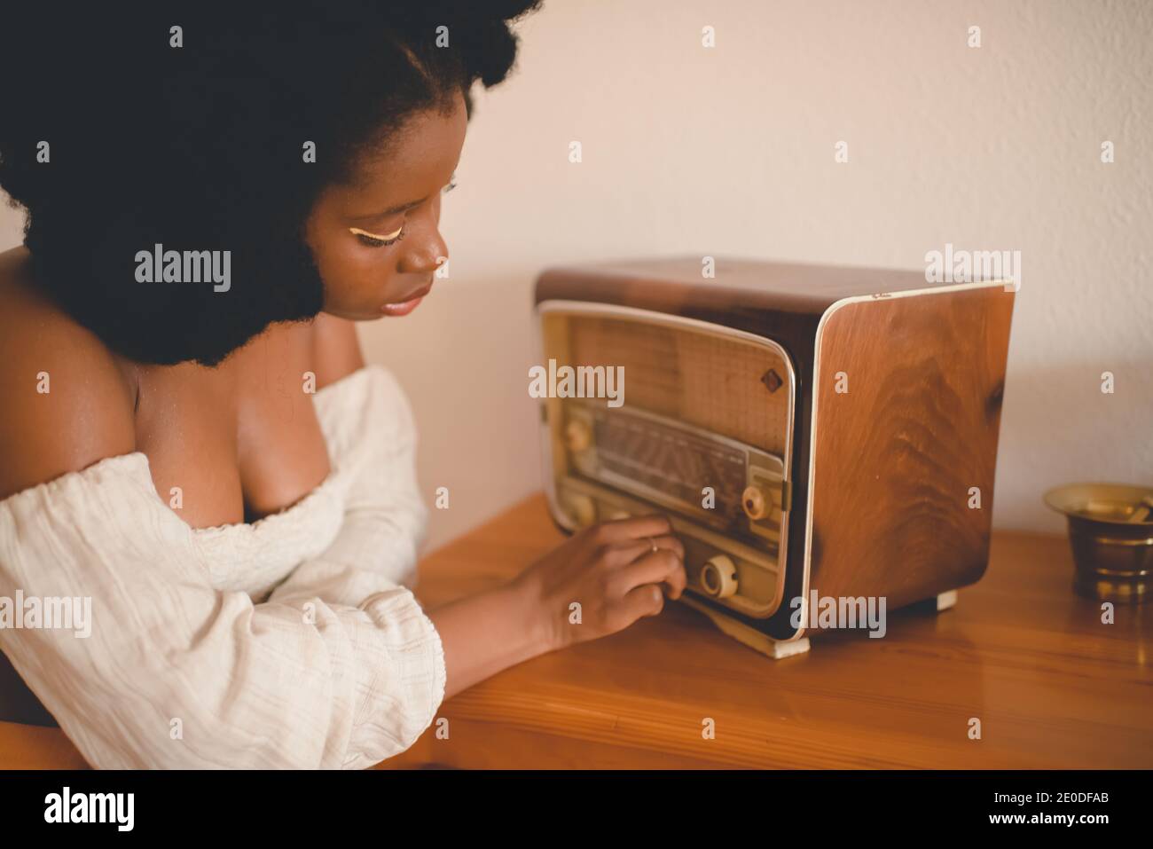 Side view of young African American female sitting at table and tuning old fashioned radio receiver while resting at home Stock Photo