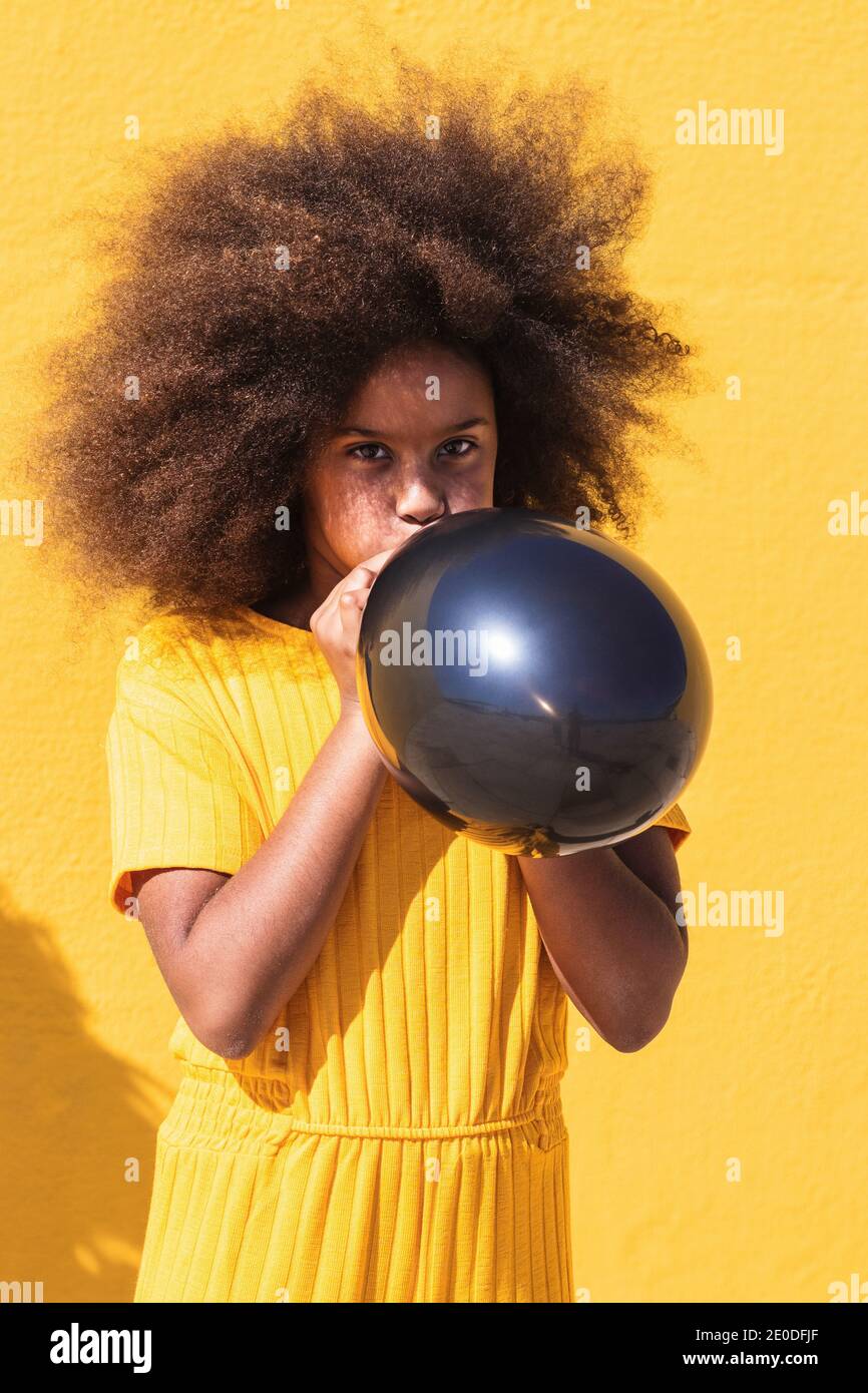 African American teen girl with curly hairstyle inflating black balloon  while standing against yellow background Stock Photo - Alamy