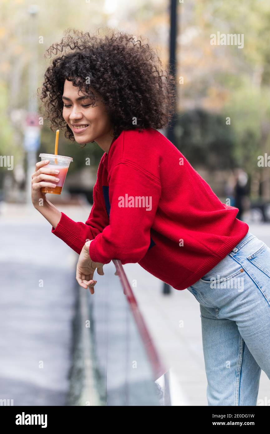 Side view of young African American female standing on street with fresh juice in plastic takeaway cup and enjoying weekend in city Stock Photo