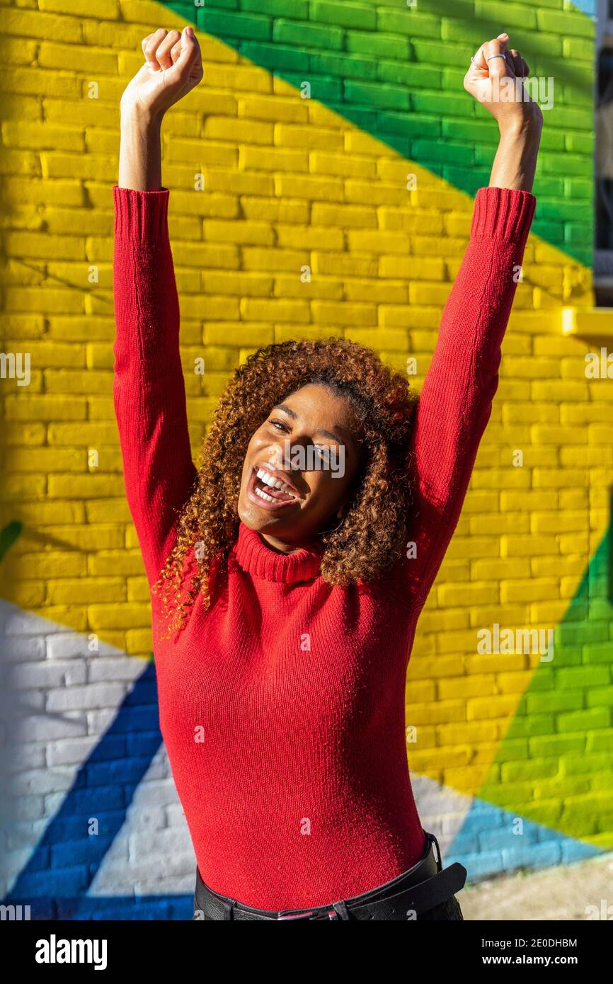 Excited young ethnic lady with long curly hair in casual clothes screaming with fists up while standing on street near colorful graffiti wall Stock Photo