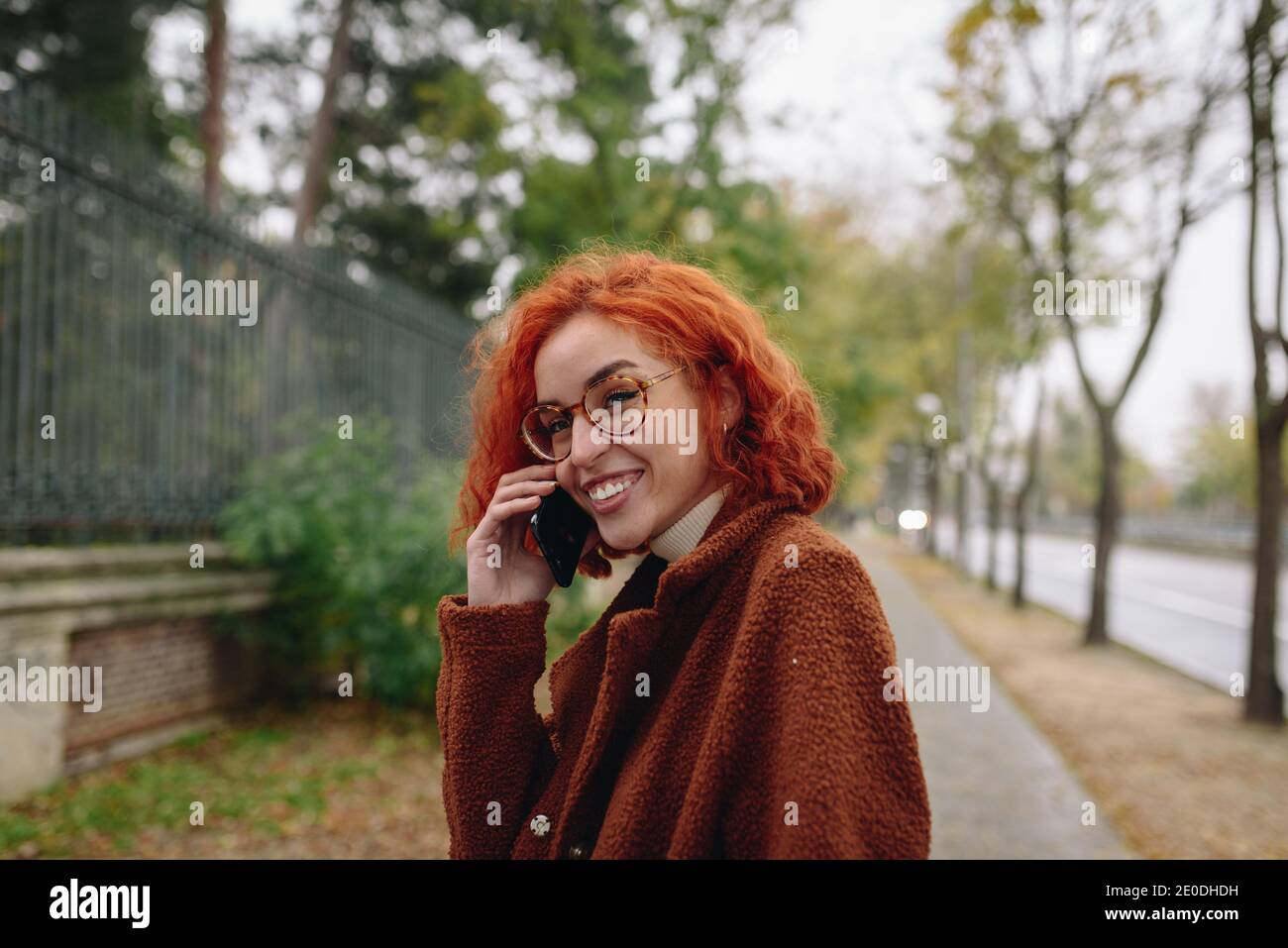 Delighted female with red hair and in autumn coat standing at the park and speaking on mobile phone while enjoying conversation Stock Photo