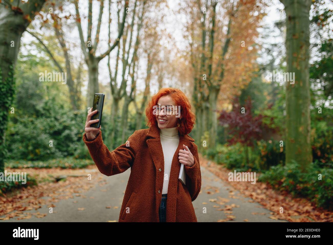 Optimistic female with red hair and in autumn coat standing on alley in park and taking selfie Stock Photo
