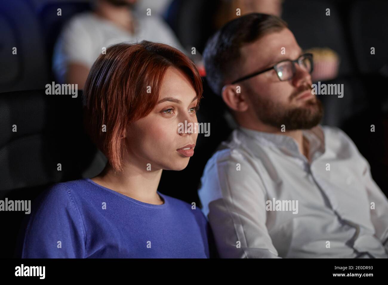 Selective focus of young couple watching movie in cinema, sitting in comfortable black seats. Side view of concentrated caucasian bearded man and woman with strong faces enjoying film. Stock Photo