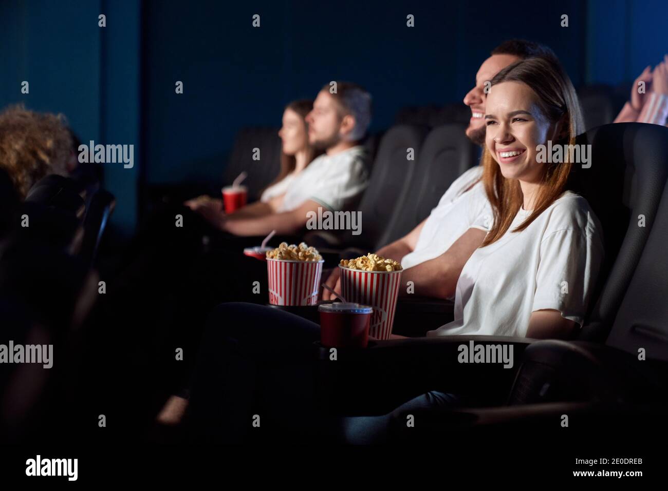 Selective focus of young girl laughing from weird situation, watching comedy in cinema. Side view of concentrated caucasian man and woman wearing white t shirts enjoying funny film. Stock Photo