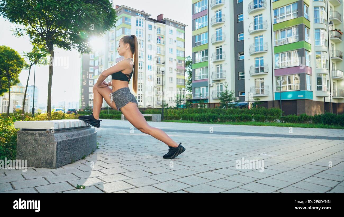 Gorgeous woman in sportswear stretching leg on bench, doing flexibility exercises after running workout in city streets. Stunning girl warming up in rays of morning sun. Healthy lifestyle concept. Stock Photo