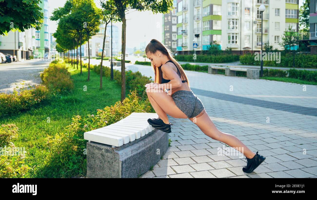 Beautiful flexible woman in sportswear stretching leg on bench, practicing flexibility exercises after running in city streets. Stunning woman warming up under rays of morning sun. Workout concept. Stock Photo