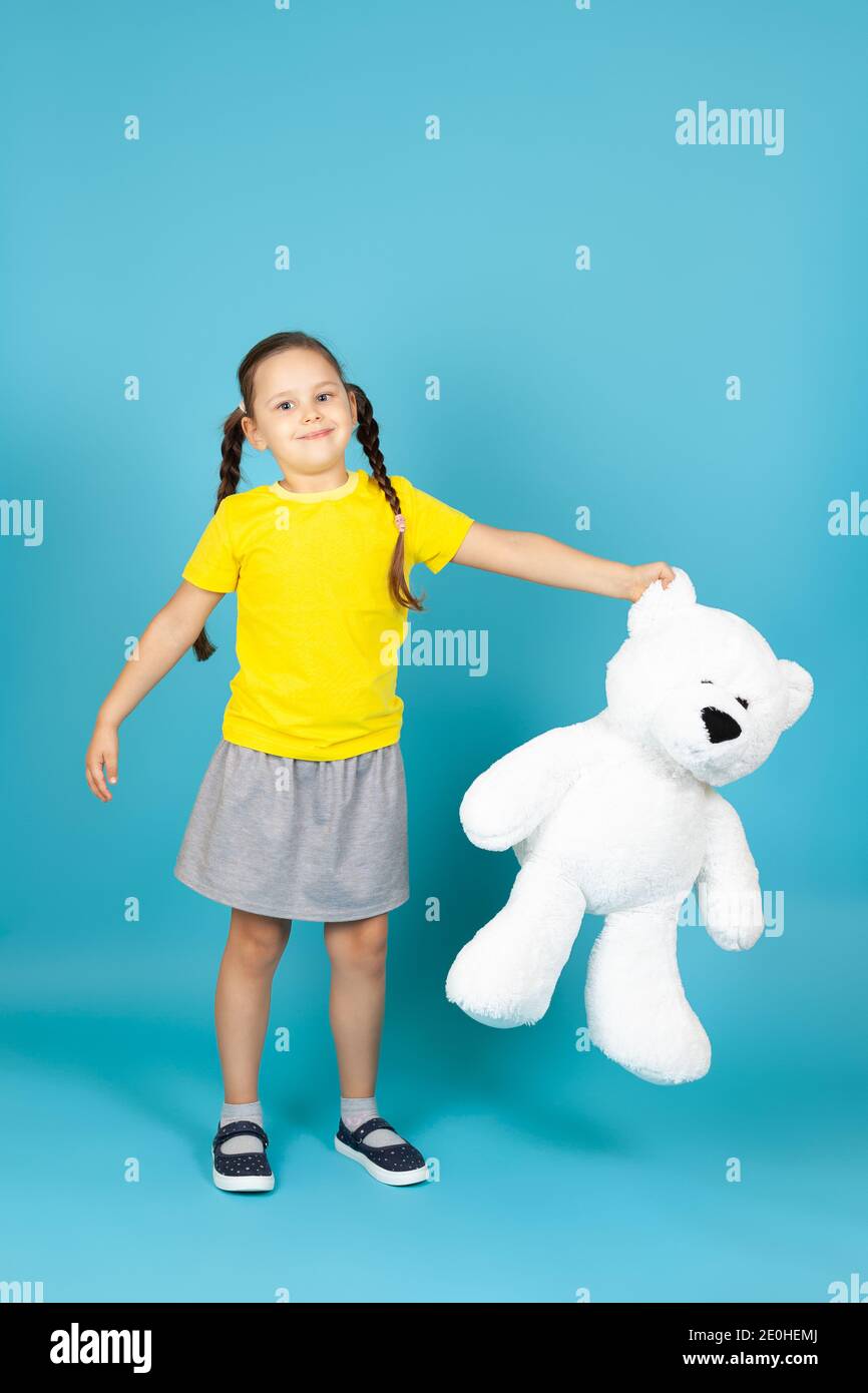 a full-length girl with pigtails in a yellow T-shirt and gray skirt holds a large white teddy bear by the ear , isolated on a blue background Stock Photo