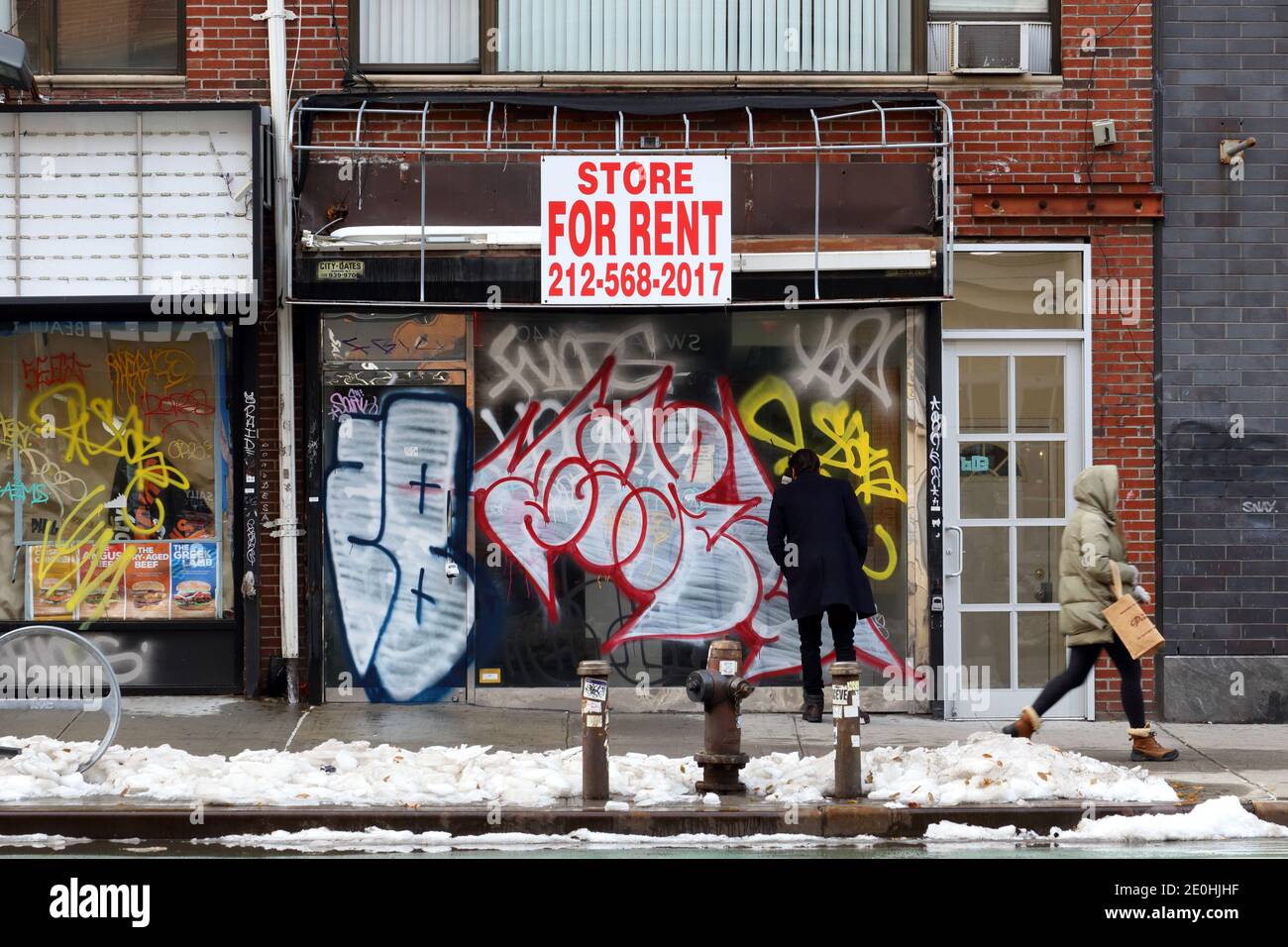 A For Rent sign hangs above a vacant storefront vandalized with graffiti with a person peering through the windows, in New York City. Stock Photo
