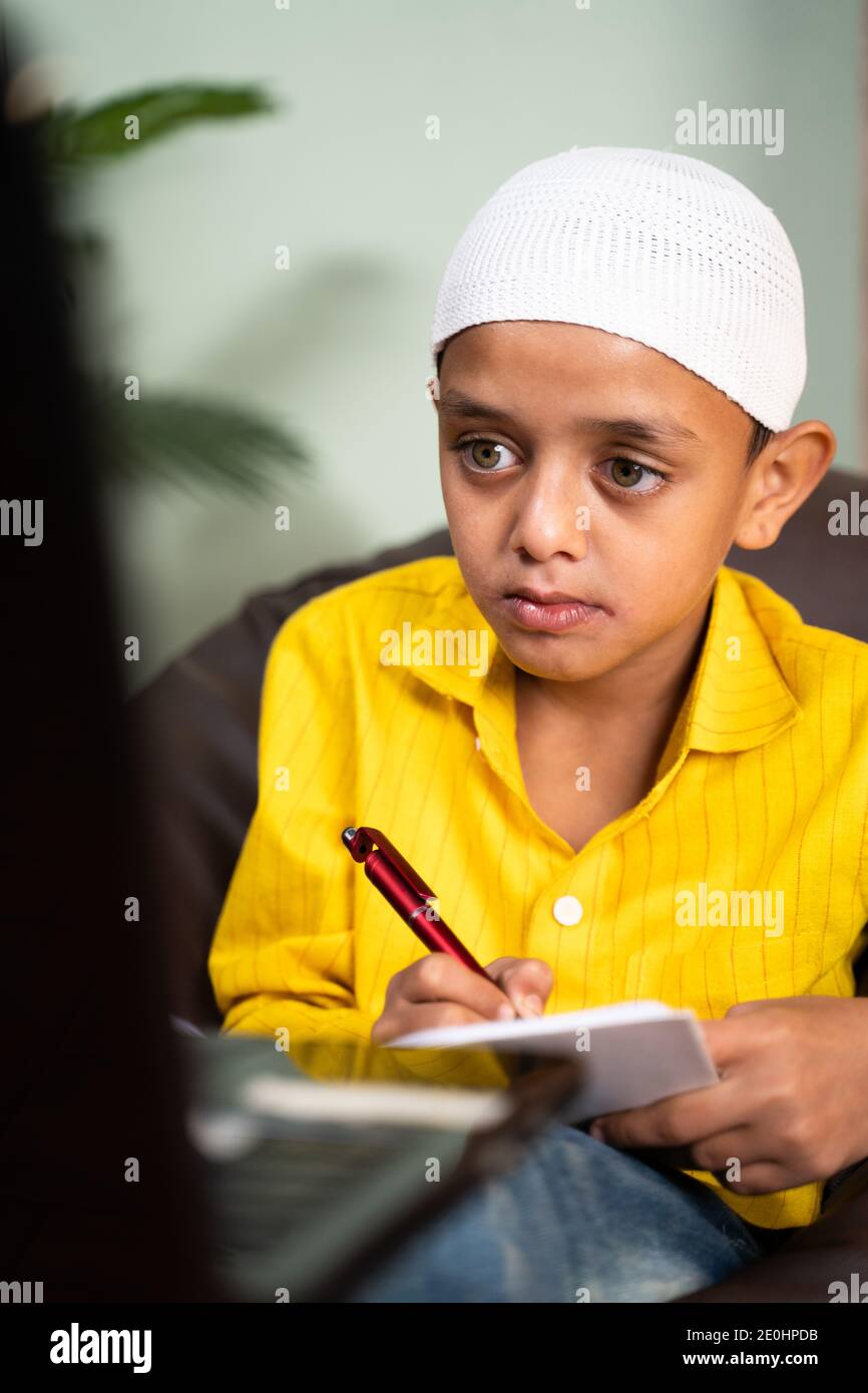 Muslim kid writing notes during online class in video call on laptop - concept of e-learning, virtual education and home schooling due to coronavirus Stock Photo