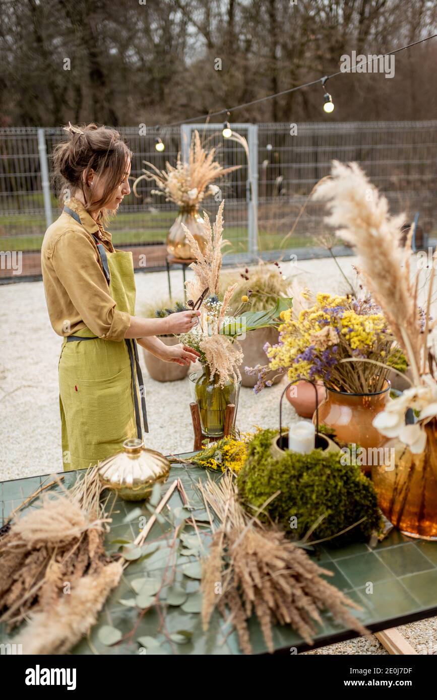 Young woman making compositions of dried and fresh flowers and herbs at the workshop outdoors. Florist, gardener or decorator composing floral decoration Stock Photo