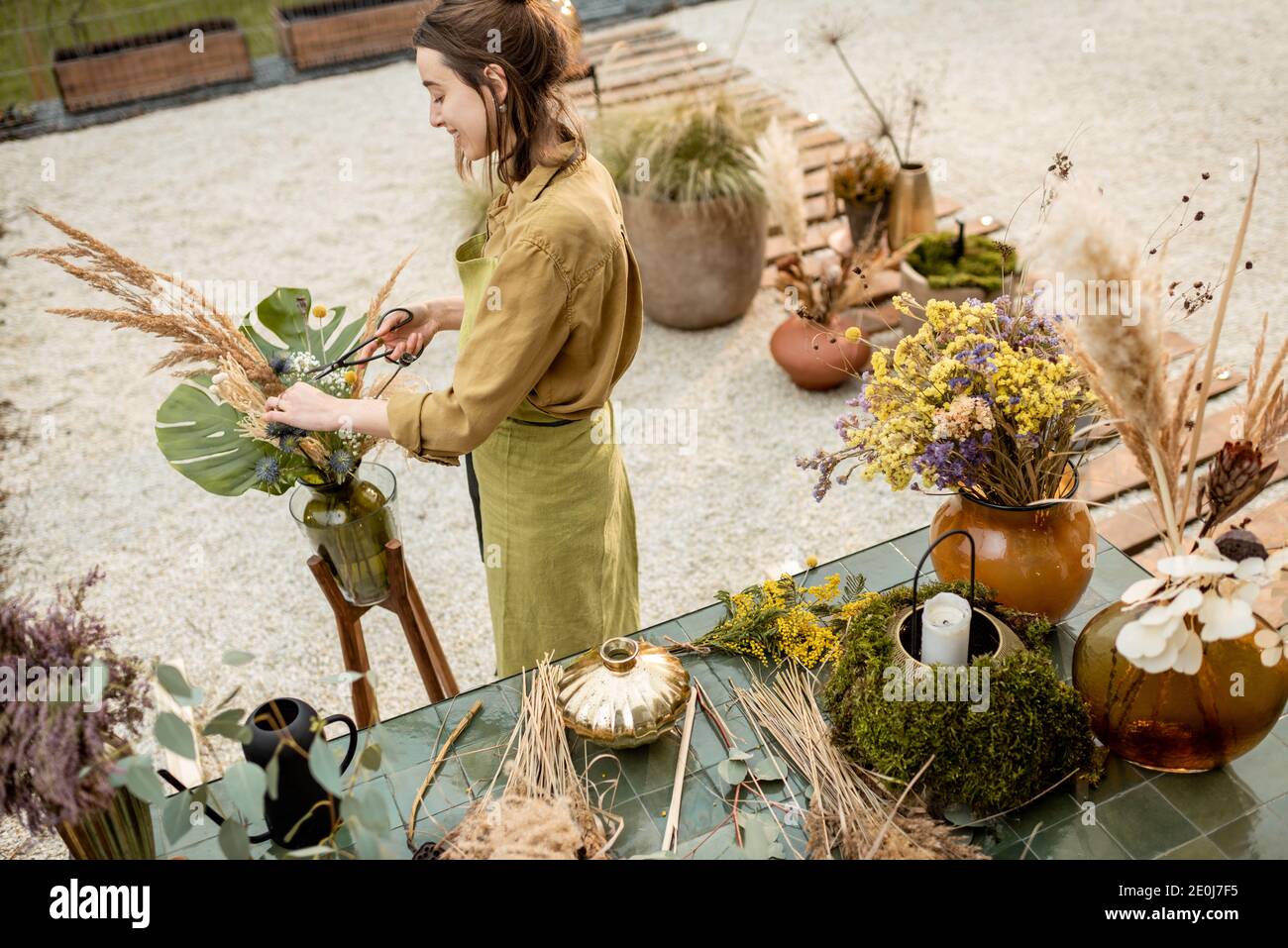 Young woman making compositions of dried and fresh flowers and herbs at the workshop outdoors. Florist, gardener or decorator composing floral decoration Stock Photo