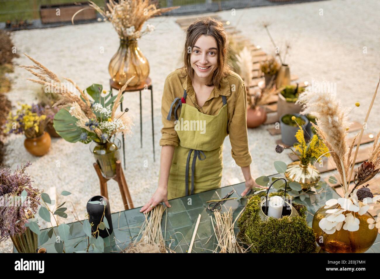 Portrait of a cheerful woman making compositions of dried and fresh flowers and herbs at the workshop outdoors. Florist, gardener or decorator profession or hobby concept Stock Photo