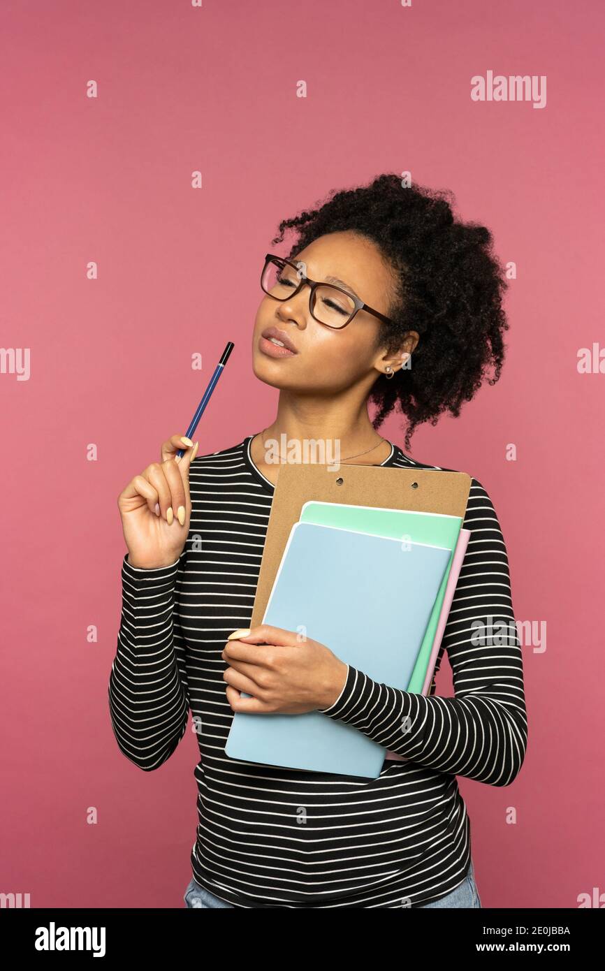 Pensive Afro-American teacher woman isolated on pink studio wall. Student girl wear glasses holding folder, notebooks and pen, thinking, looking away. Stock Photo