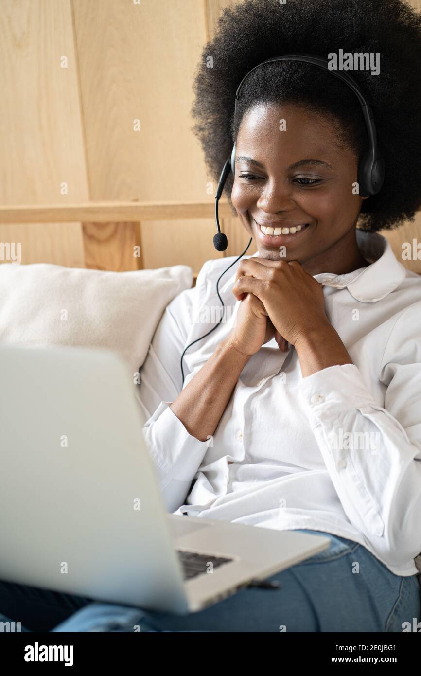 Smiling Afro-American millennial woman wearing headset, communicating via video chat on laptop, talking in zoom, watching webinar or video stream Stock Photo