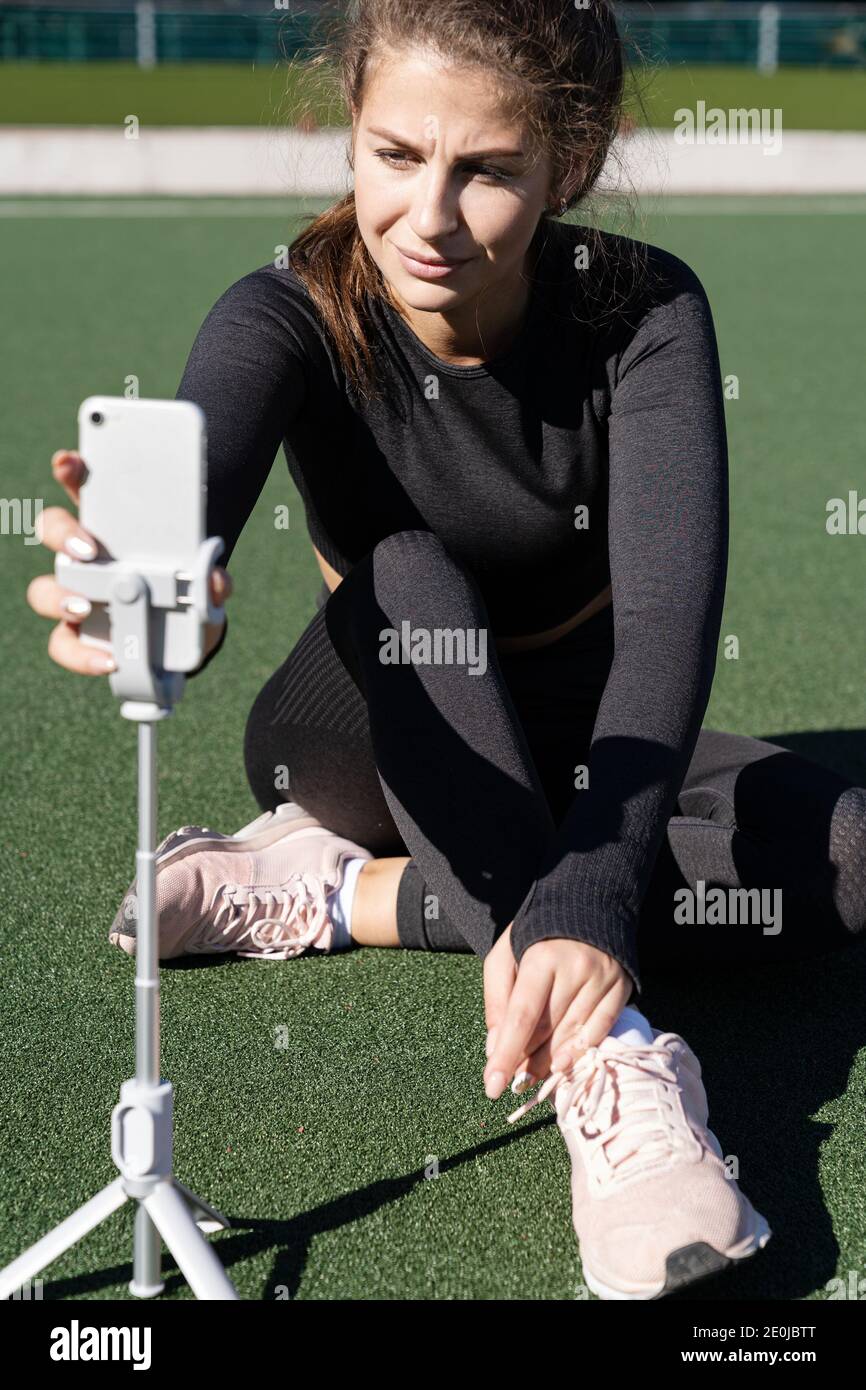 Woman in sportswear vlogger sitting on artificial turf outdoor, demonstrating exercises for her online blog, starting recording on smartphone camera o Stock Photo