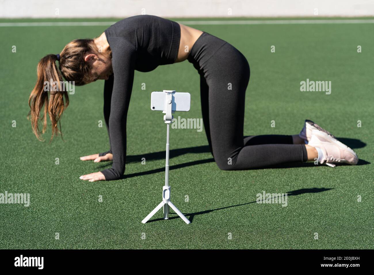 Woman doing yoga exercise called Cat pose outdoor, demonstrating exercises for her online blog, records on smartphone camera on tripod. Streaming a le Stock Photo