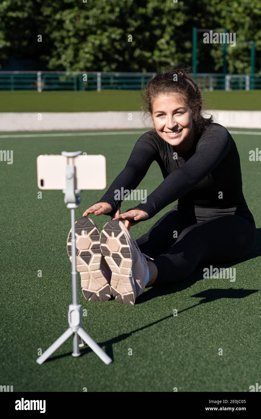 Smiling woman vlogger in sportswear sitting on artificial turf outdoor, demonstrating exercises for her online blog, records on smartphone camera on t Stock Photo