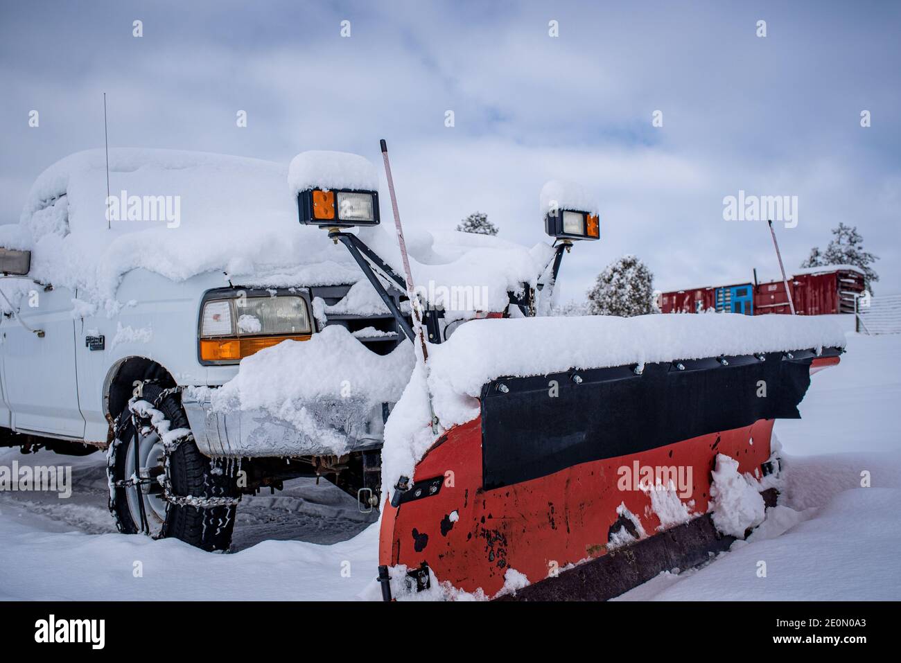 A white truck with a red snow plow and add on lights parked in the snow in front of a traincar