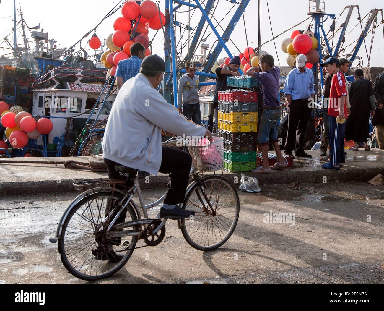 A man on a bicycle watches fisherman packing plastic containers with fish and ice at the port of Essaouira in Morocco in the late afternoon. Stock Photo