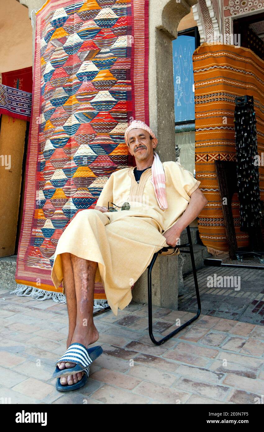 A man sits outside his carpet shop in Meknes, Morocco. Meknes is named after a Berber tribe which, was known as Miknasa, native Berber name: Imeknasen Stock Photo
