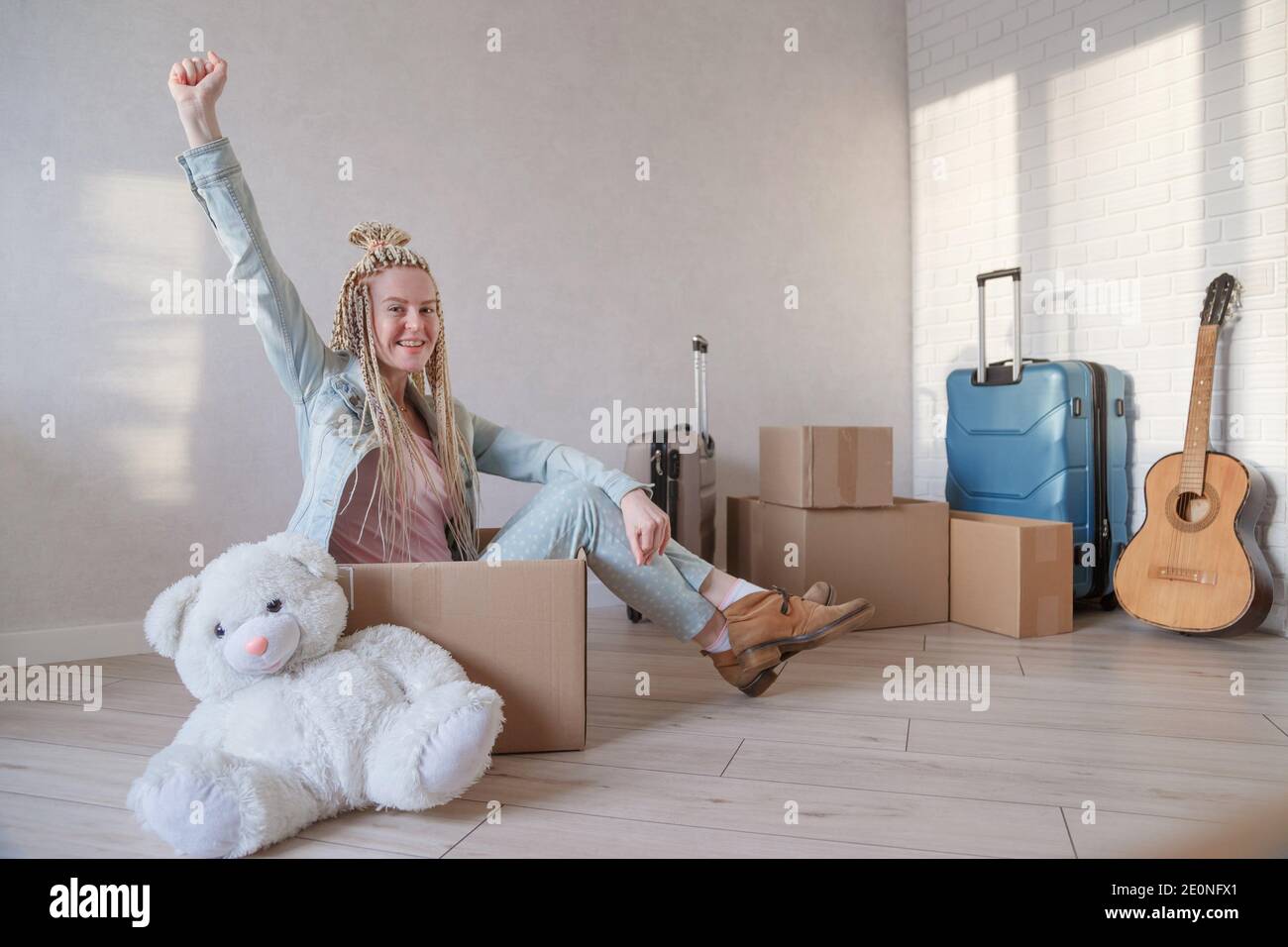 Young woman with pigtails is happy to move to new flat. Sits in a box with raised hand. Packed luggage and belongings and guitar on the background. Stock Photo