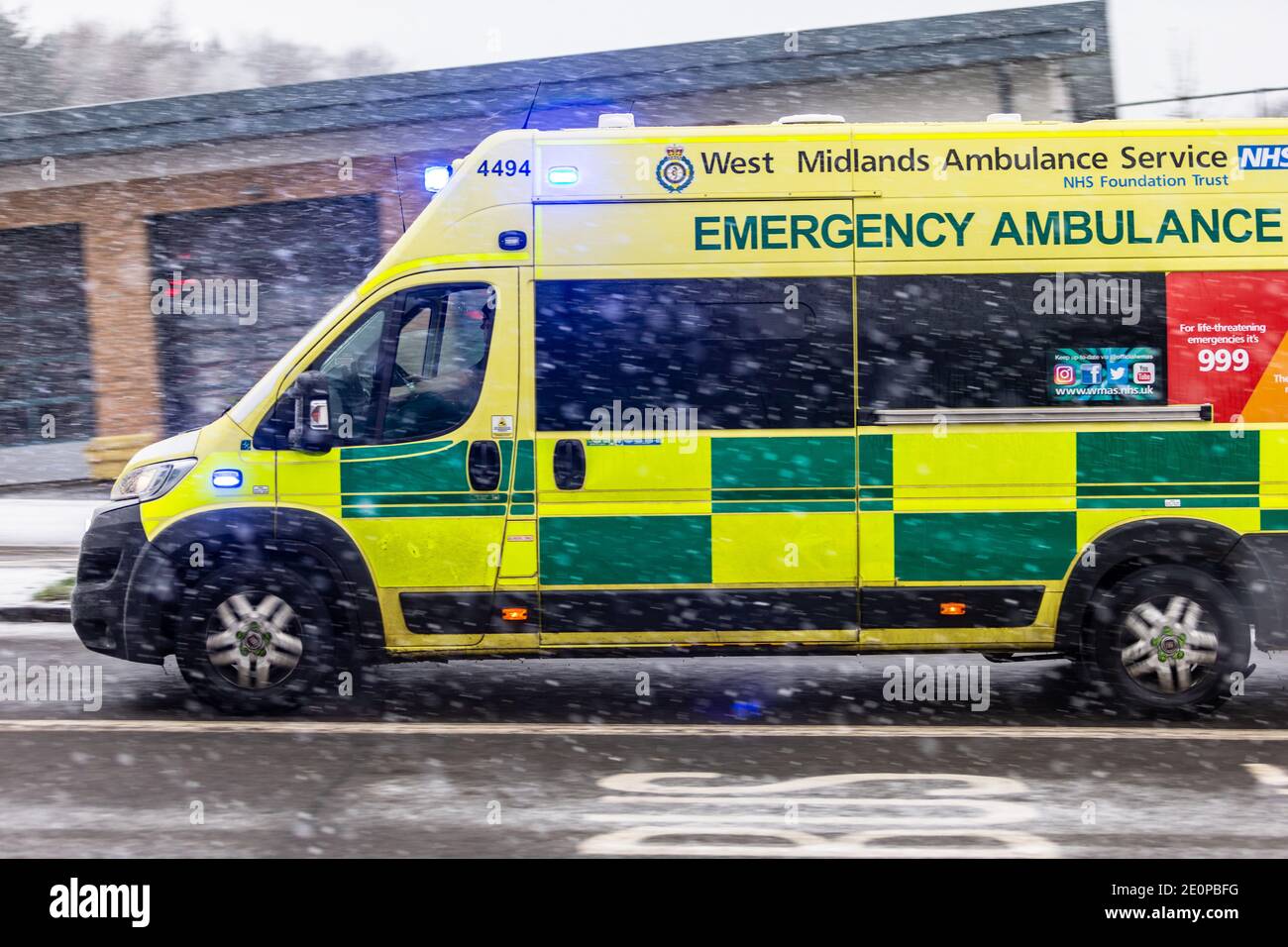 An emergency ambulance makes its way through falling snow in a town, UK Stock Photo