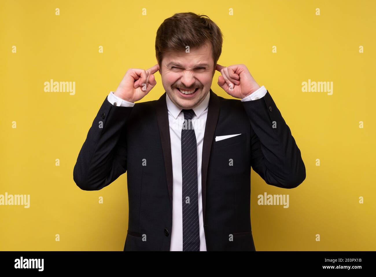 This is too loud. Frustrated young man in formalwear plugging ears with his fingers and keeping eyes closed. Studio shot on yellow wall. Stock Photo