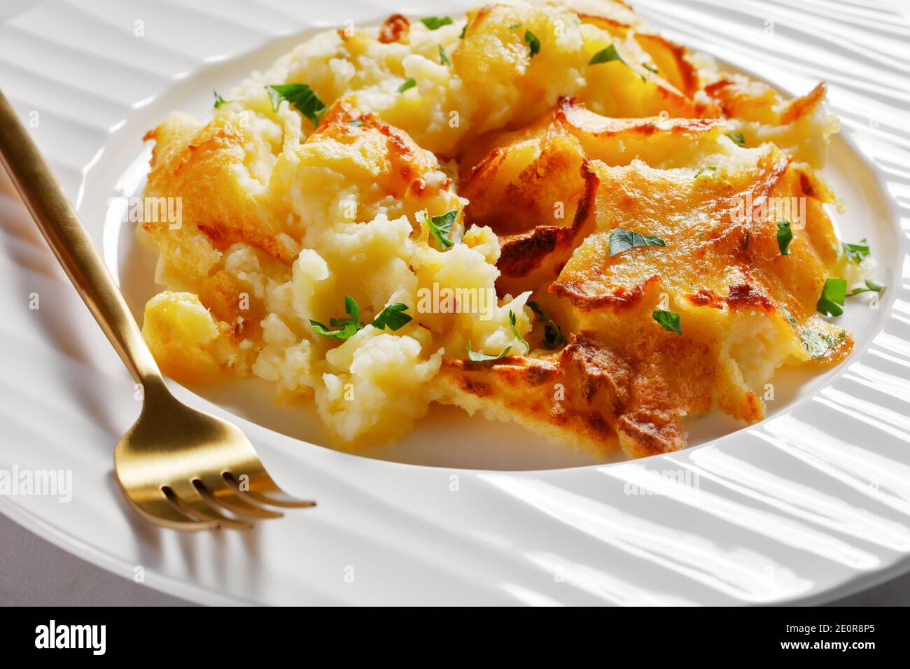 close-up of a portion of Garlic Parmesan Mashed Potato Casserole sprinkled with parsley on a white plate with golden fork, on a marble table, horizont Stock Photo
