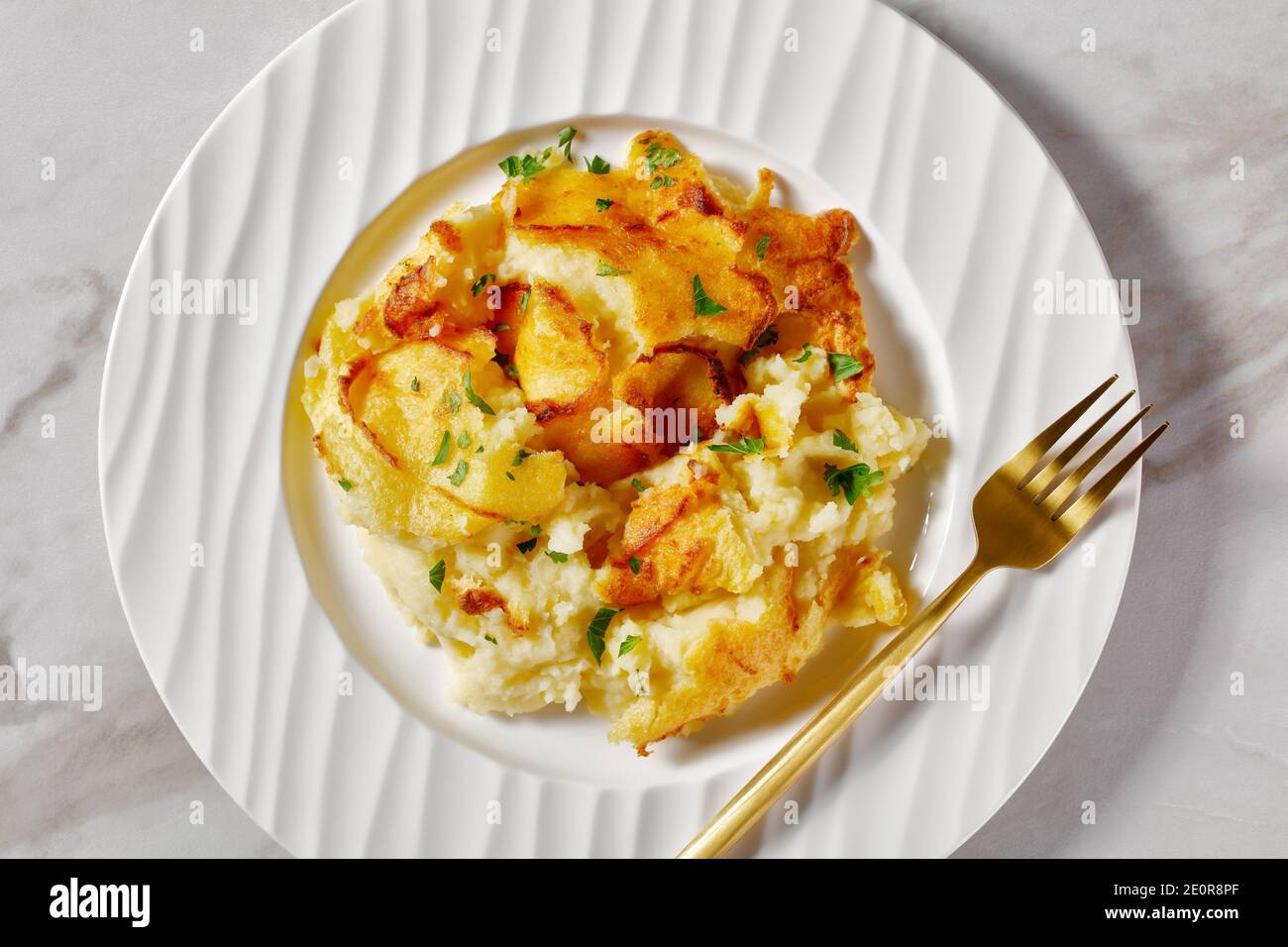 portion of baked Mashed Potato Casserole sprinkled with parsley on a white plate with golden fork, on a marble table, horizontal view from above Stock Photo