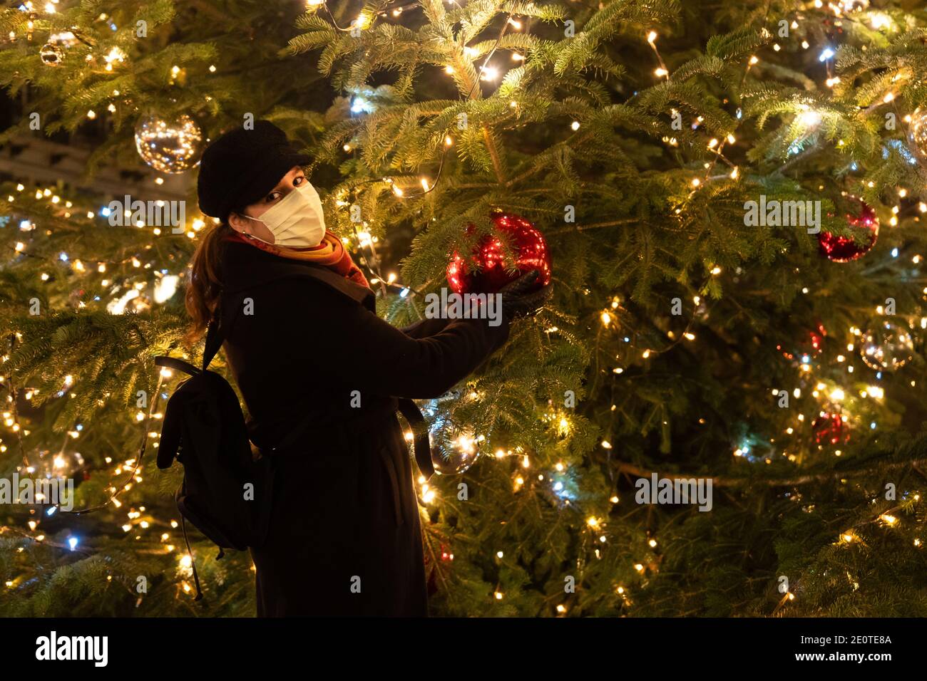 Woman holding Christmas giant red bauble. Woman with mask stands in front of big Christmas tree. Looking to camera. Stock Photo