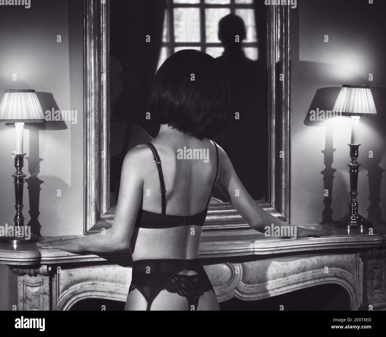 Woman with short black hair standing half naked by mantelpiece, wearing sexy lingerie, garter belt and stockings. View of her back with a shadowy dark Stock Photo