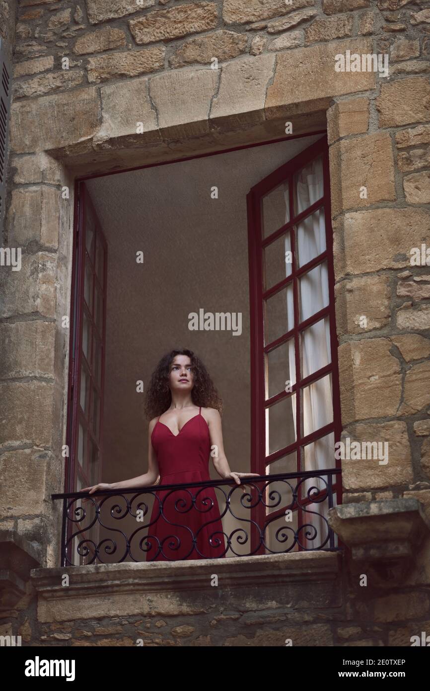 Beautiful young woman in an elegant burgundy red dress standing in an open balcony window of a medieval French stone house, looking outside. View from Stock Photo