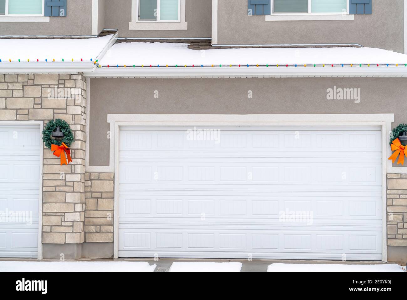 White Garage Door Of Home With Wreath And Snowy Roof Lined With Christmas Lights 2E0YK0J 