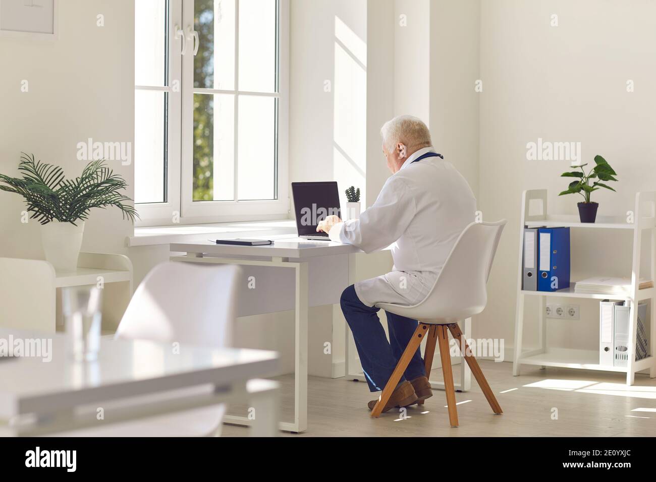 Elderly gray haired doctor in white uniform sitting at laptop and communicating with patient online during videocall in medical clinic office, rear vi Stock Photo
