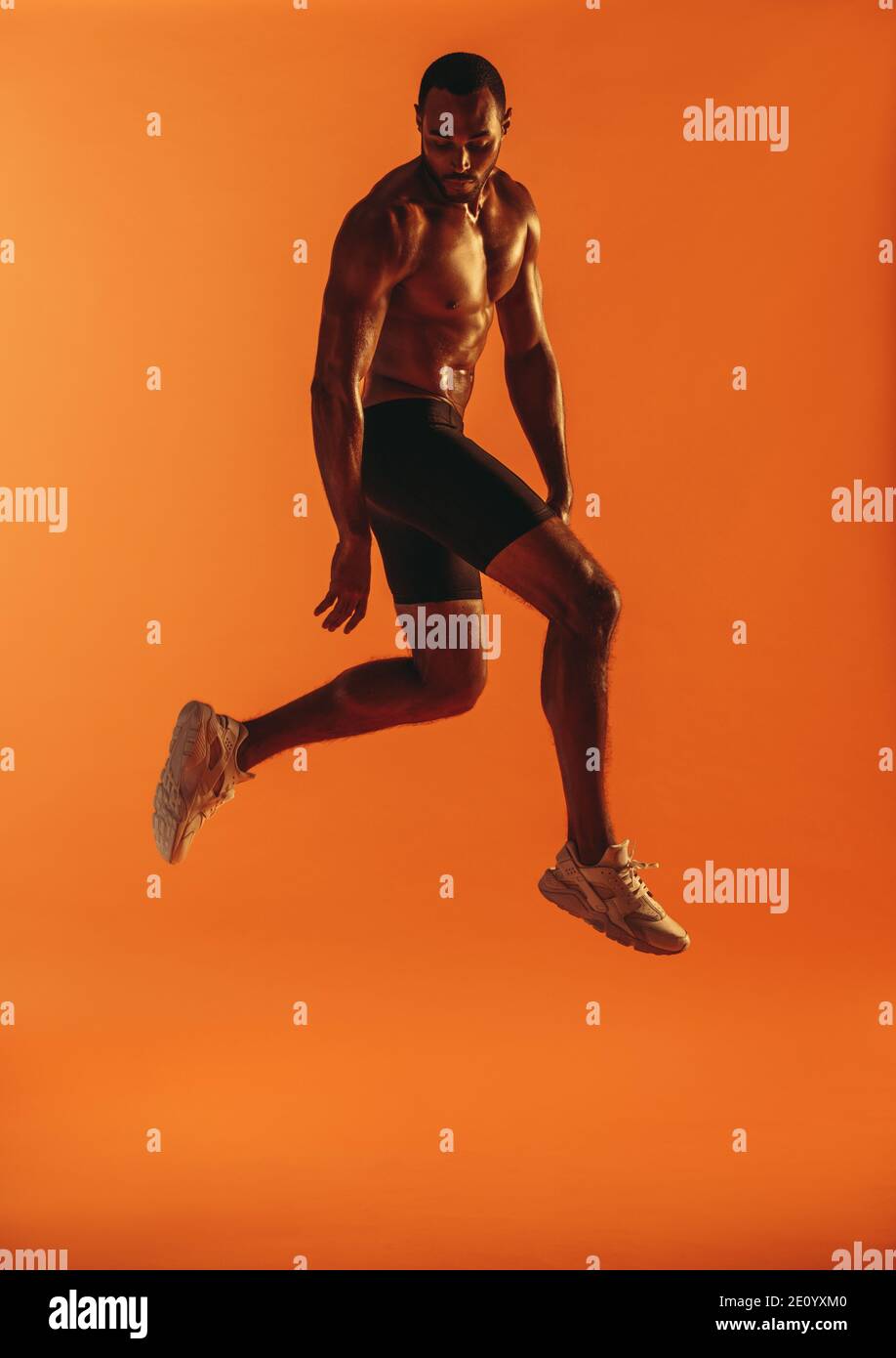 Athletic man jumping in air while working out on orange background. Monochrome fitness portrait of muscular athlete doing workout. Stock Photo