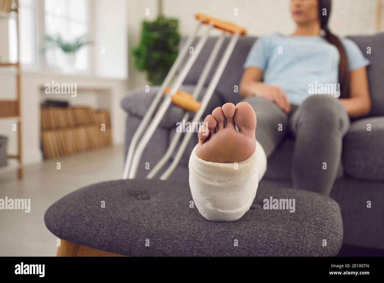 Woman with leg in plaster cast sitting on sofa at home with pair of crutches beside Stock Photo