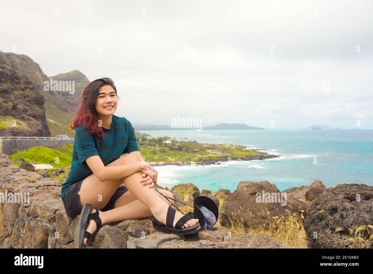 Biracial Asian Caucasian teen girl sitting on top of rock stone wall at Makapu'u Lookout looking out over blue Pacific ocean on Oahu island with mount Stock Photo