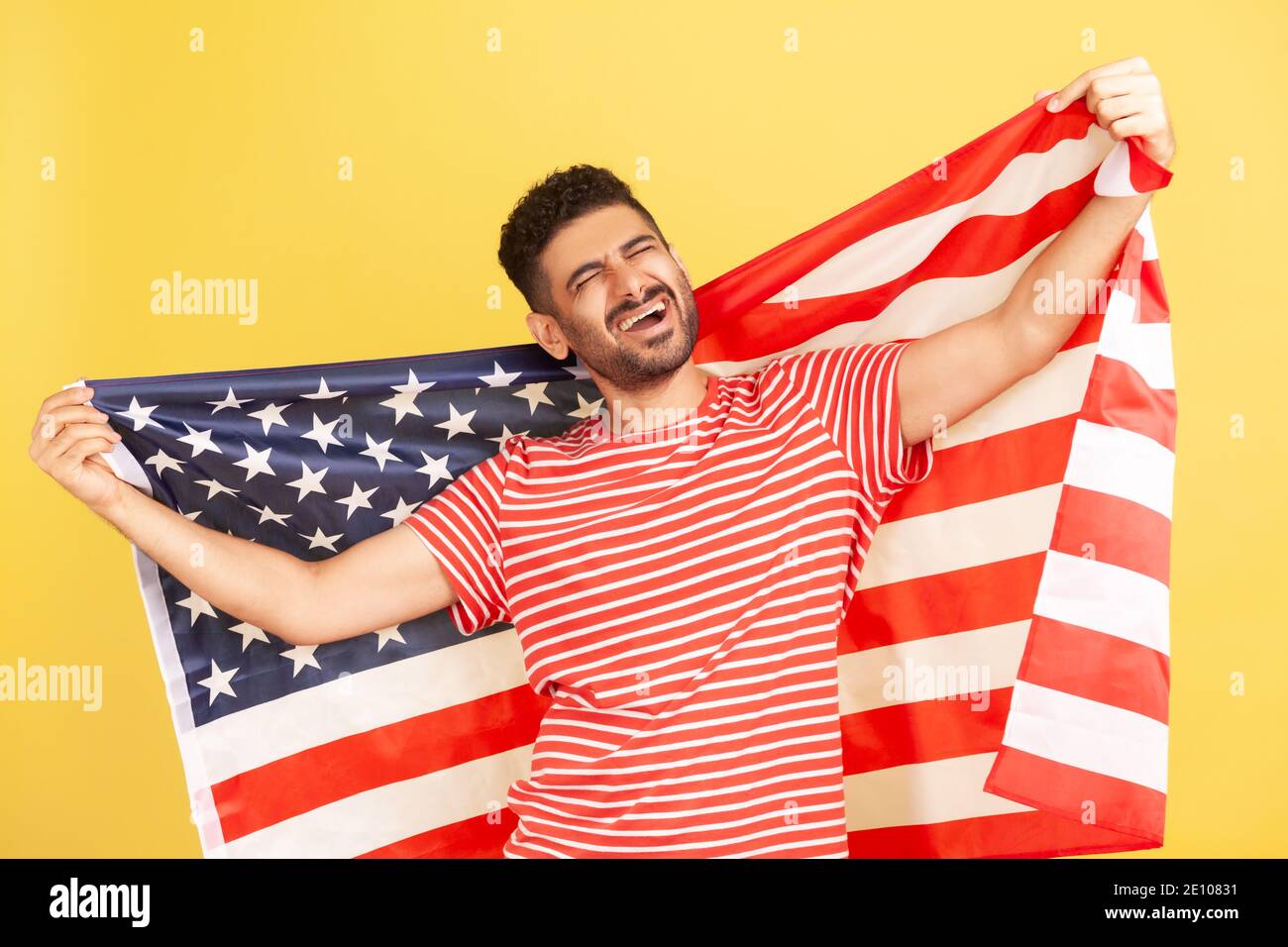 Excited patriotic man with beard in striped t-shirt standing holding in hands flag of united states of america and screaming, singing anthem. Indoor s Stock Photo
