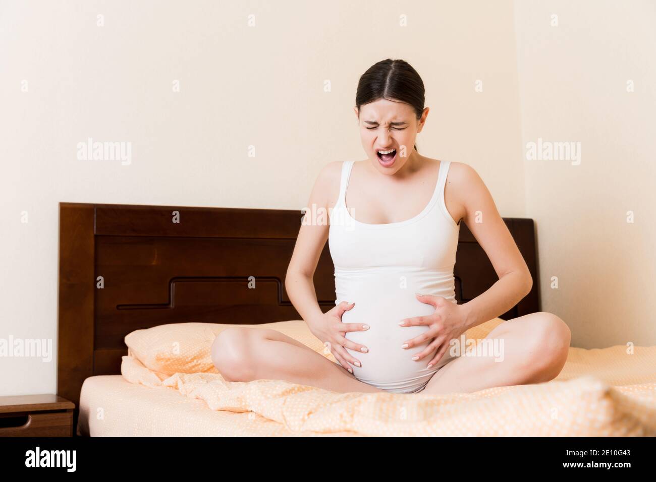 Young pretty pregnant woman, in pain, sitting on bed in bedroom