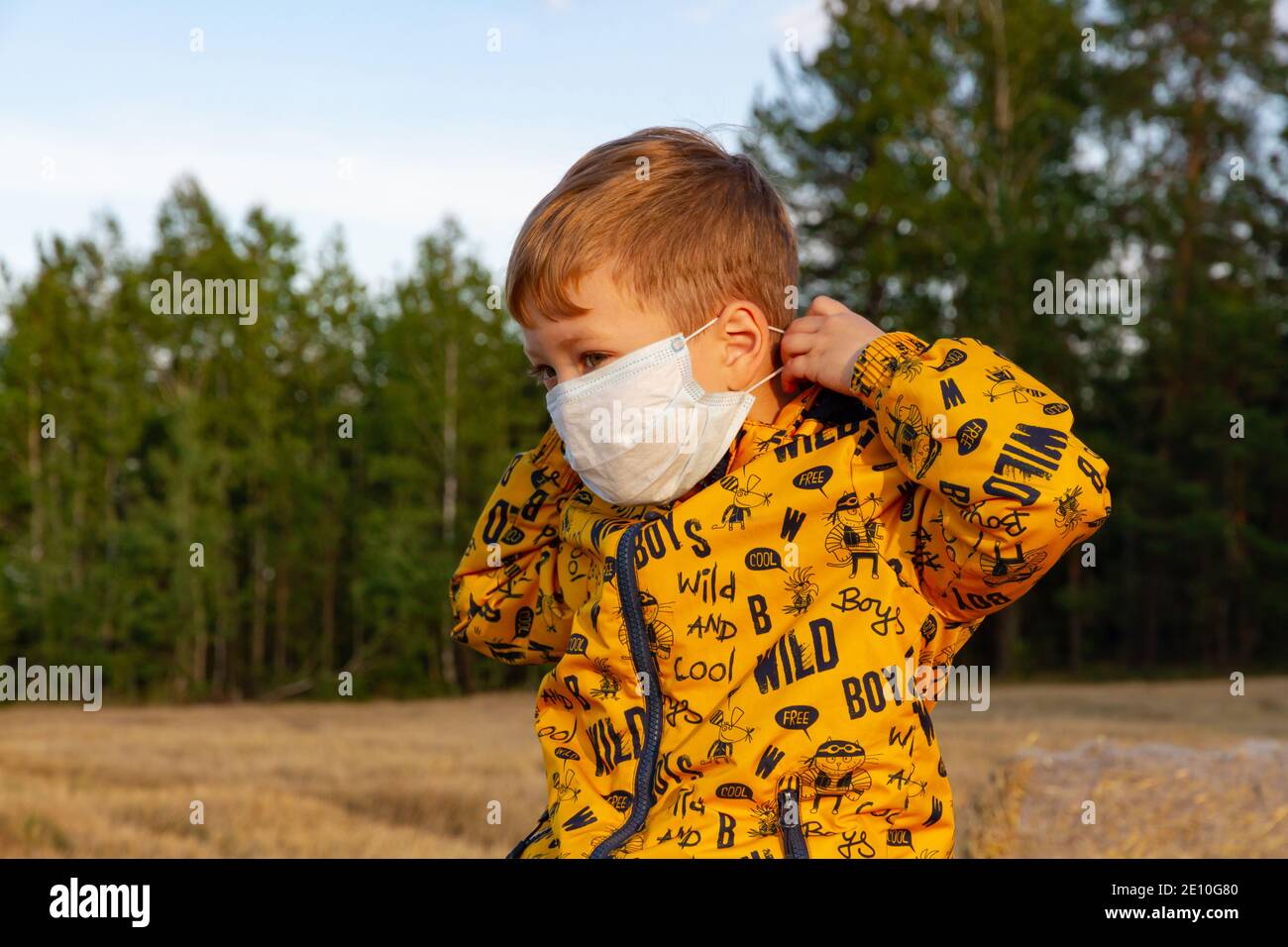 a boy in a yellow jacket puts on a medical mask on a bright sunny day Stock Photo