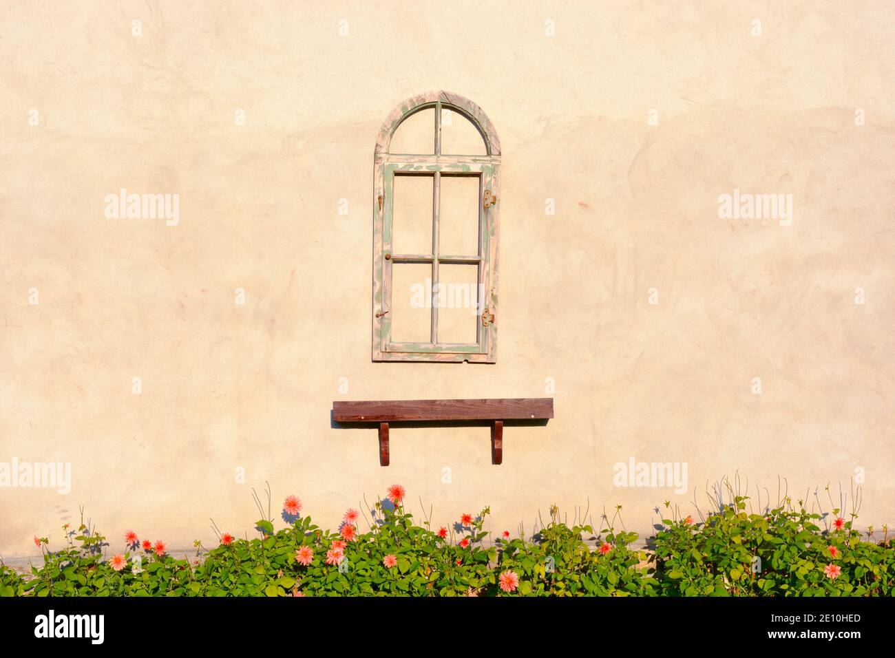 The rustic wooden window frame and the brown shelf below hang on the empty house wall. Below a flowerbed with blooming pink flowers. Stock Photo