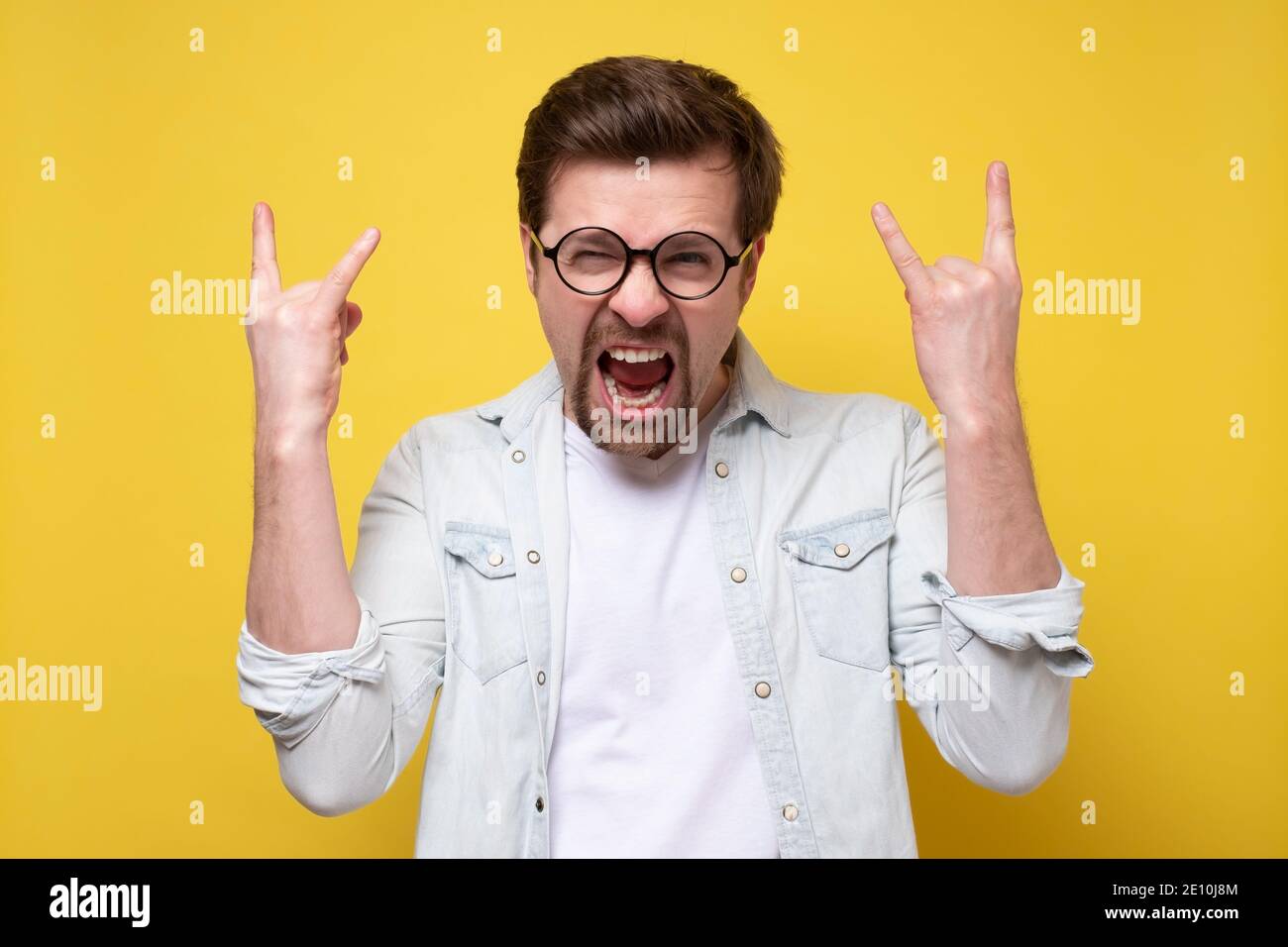 Caucasian man showing rock signs with both hands and shouting out loud, expressing overwhelmingness and joy. Studio shot on yellow wall. Stock Photo