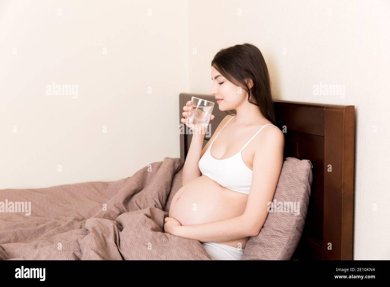 Photo of pretty pregnant woman keeping her hands on belly while drinking water on bed at home. Stock Photo