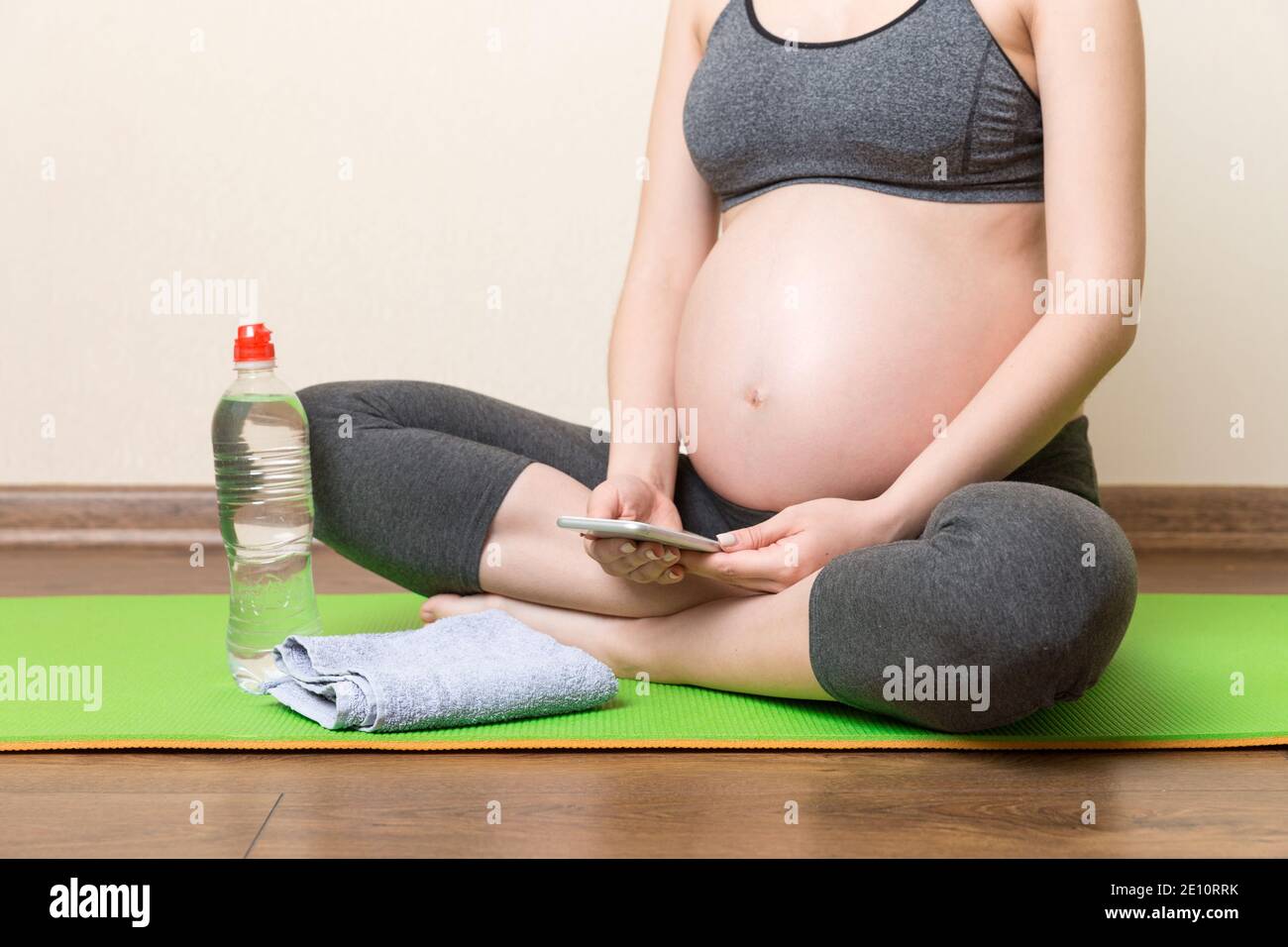 Pregnant female holding smartphone while sitting on exercise yoga mat at home at coronavirus time. Stock Photo