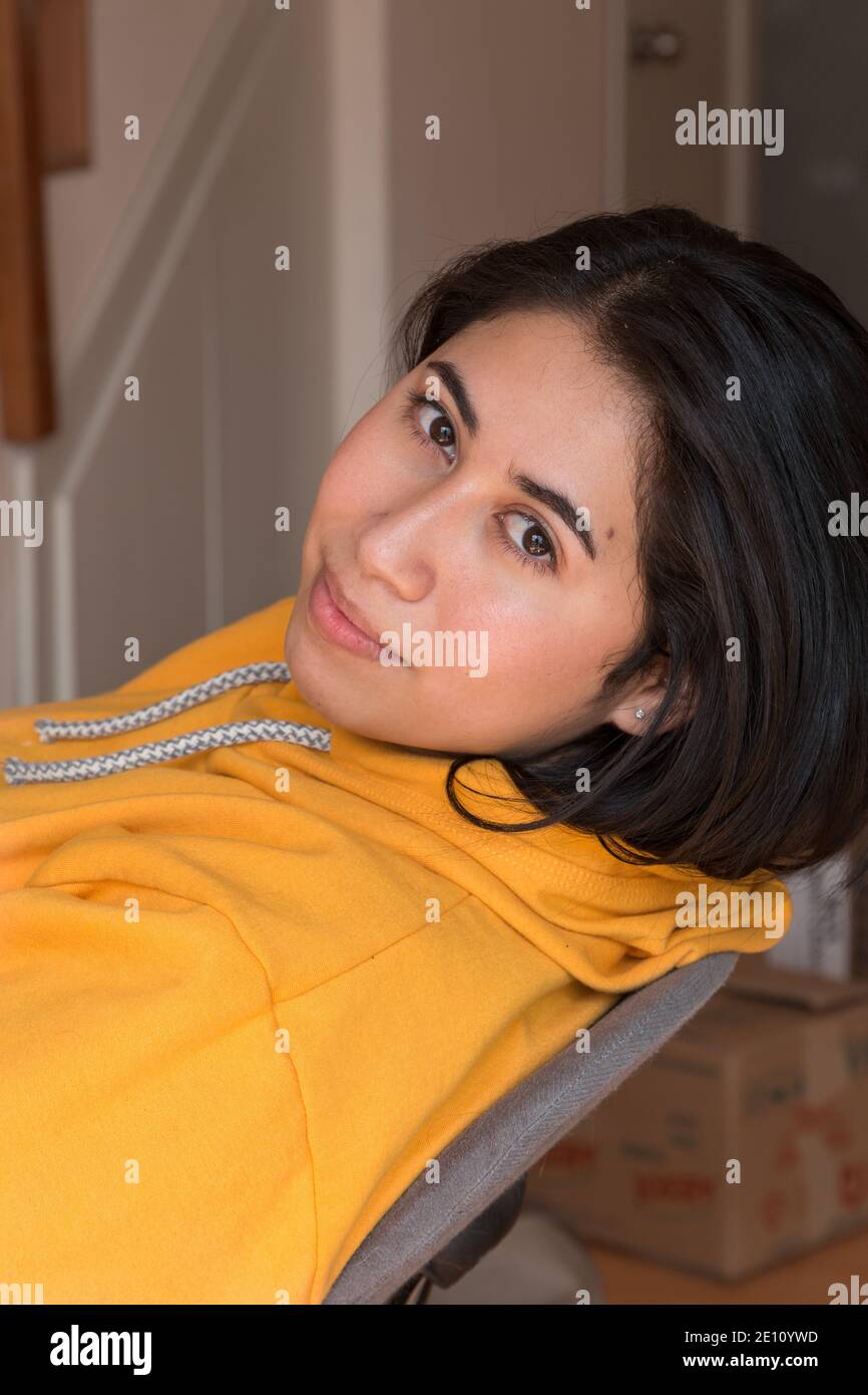 Latin young woman with a yellow sweatshirt, girl sitting on a chair very relaxed inside the house on a sunny afternoon inside the house Stock Photo