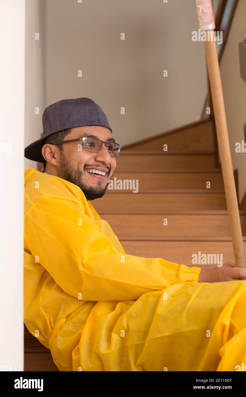 worker man in yellow rainwear sitting on the edge of some wooden bleachers with cap, glasses beard and smile on a bright day Stock Photo