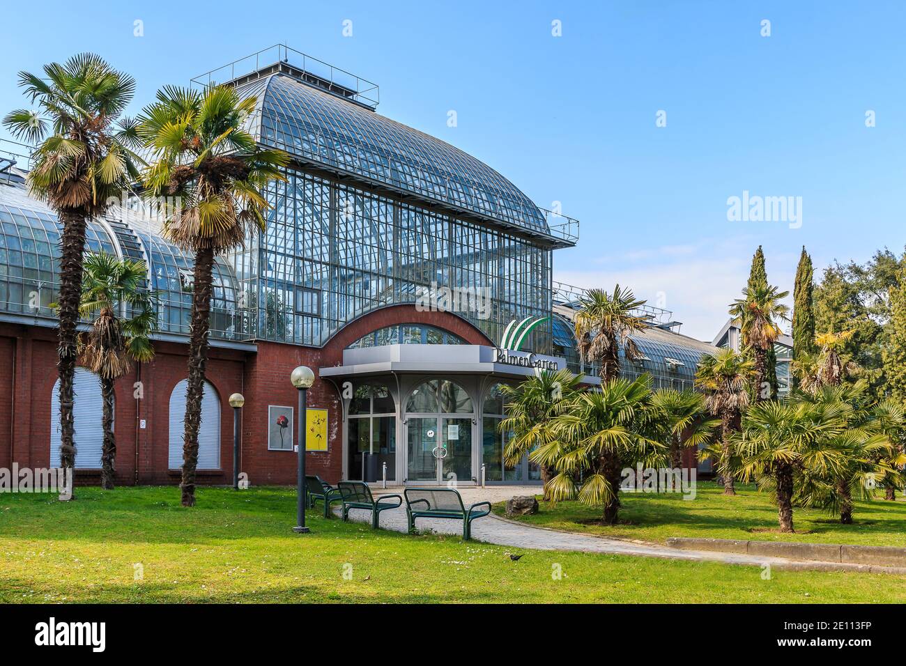Buildings of palm garden in Frankfurt. Palm trees with meadow in front of the entrance. Glass roof with windows and bricks from the building. Path wit Stock Photo