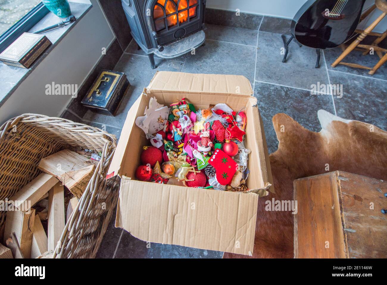 Brown cardboard box with a jumble of family colourful Christmas tree decorations infront of a log burner on a stone floor being packed away Stock Photo